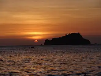 Photo of a sunsetting on the horizon of a body of water. The sky is orange and yellow, a silhouette of an island is visible, and the water is dark blue.