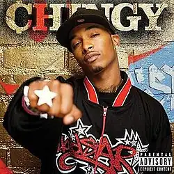 An image of a man standing in front of a gold brick wall with graffiti and the artist's name, wearing a black hoodie with the album title colored in red, and holding his right hand as a fist that shows a star ring.