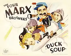 Four disheveled men with goofy expressions wearing antiquated military garb ride a rocket. One of them—with round-rim glasses, thick eyebrows, and a thick mustache—holds a telescope.