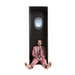 A plain white background. Mac in a gray suit and tie, barefoot, sitting down inside a locker. The inside of the locker has a window on the inside, with a blue sky behind it.