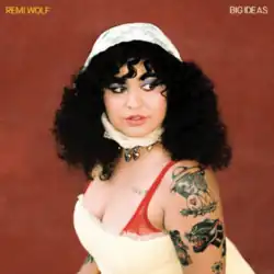 Wolf, with shoulder-length curly black hair, wearing a white dress and red bra, looking to the side in front of a brown wall.