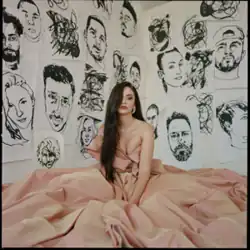 A woman's salmon-coloured dress covers the floor she sits on. Her long dark hair covers her right eye. Ink portrait drawings cover the walls behind.
