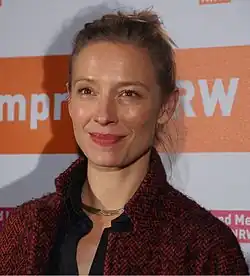 Sandra Borgmann wearing a black, open-necked top under a black and red wool coat, standing on an event stage, smiling, and looking just right of camera