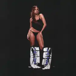 McRae standing against a black background wearing hockey leg pads with the album title on them.