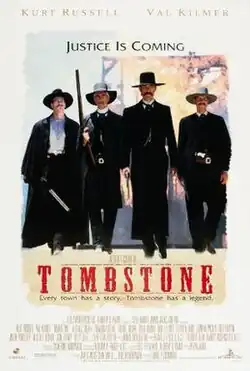 Four men dressed in black armed with pistols and rifles walking down a dusty street